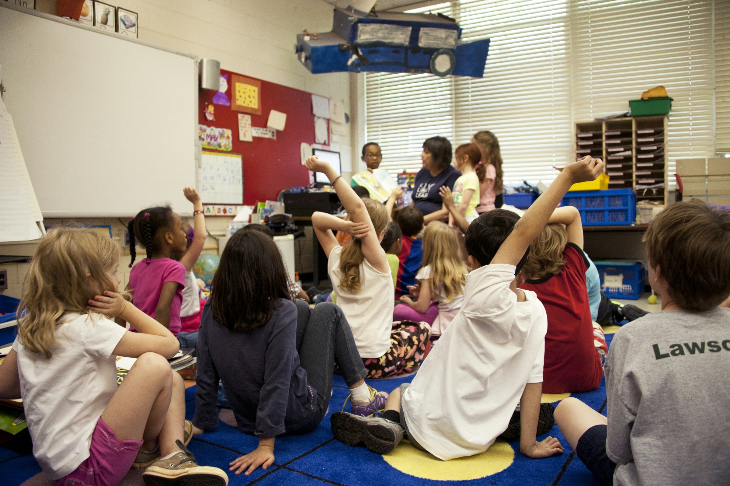 Children on a classroom carpet with hands raised